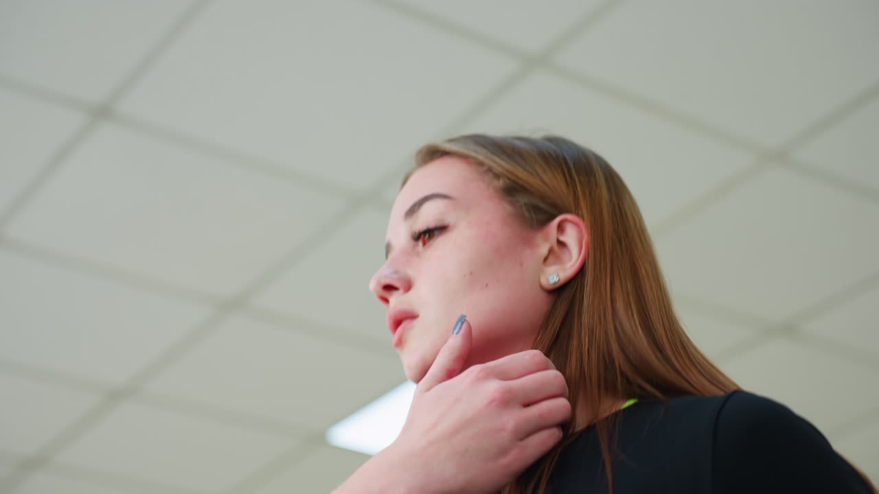 upward angle of young woman jogging indoors wearing black outfit as she raises hand to adjust hair under bright gym ceiling lights showcasing fitness routine in motion with focused expression
