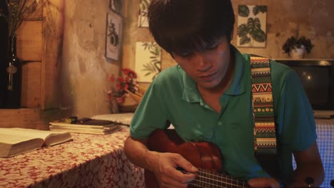 Young-Asian-male-playing-a-ukulele-while-on-a-chair-in-a-rustic-room