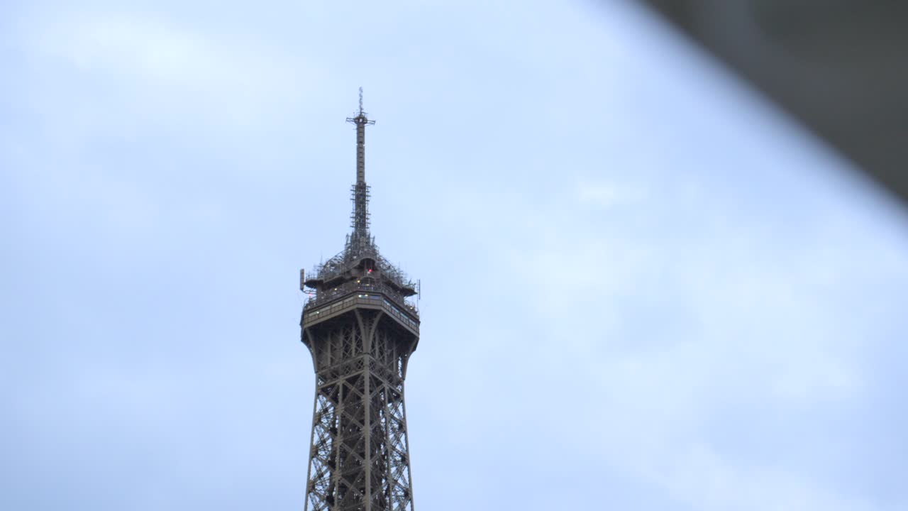 A long-lens slow-motion shot focused on the Eiffel Tower’s top floor and summit, framed against a grey overcast sky, capturing Paris’s iconic landmark in winter light