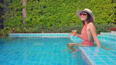 a beautiful woman in a coral bathing suit, sunglasses, and straw sun hat sits on the step of a pool while drinking a colorful tropical drink