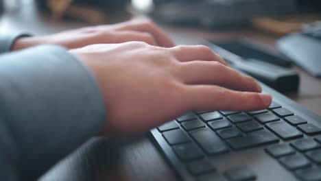 Hombre-Con-Una-Camisa-Azul,-Está-Trabajando-En-Una-Computadora-Con-Un-Teclado-Gris-Y-Un-Escritorio-De-Madera