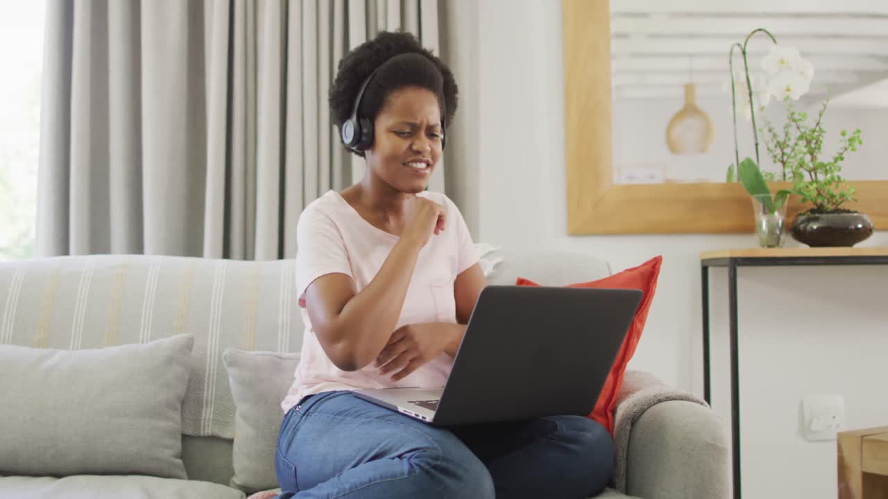 Happy african american woman wearing headphones sitting on sofa, using laptop
