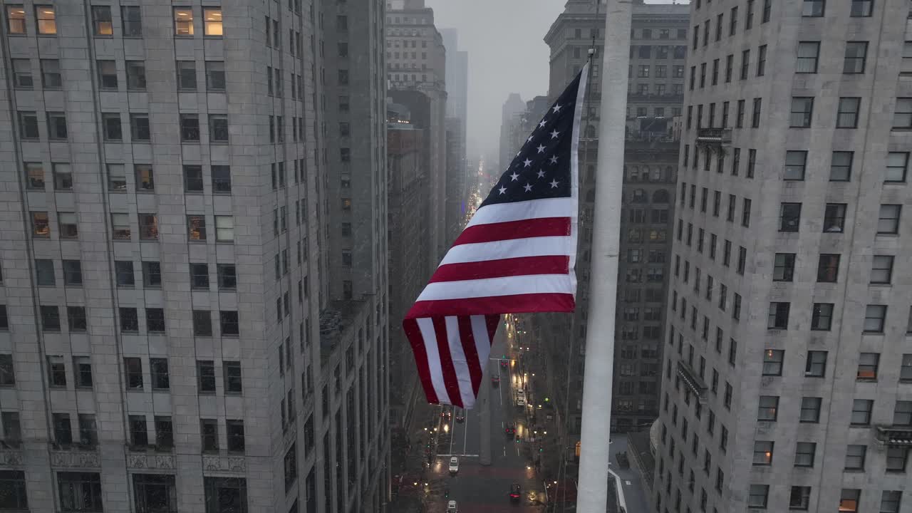 Vista aerea di una bandiera americana che sventola tra alti edifici del centro, mentre i semaferi brillano sotto durante un clima nevoso e piovoso in una fitta strada urbana. Ripresa cinematografica con drone. Stato patriottico.