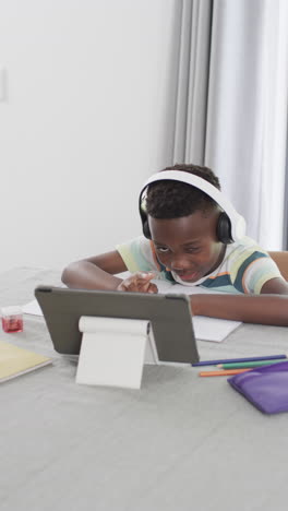 vertical video: african american boy wearing headphones, looking at tablet and doing homework