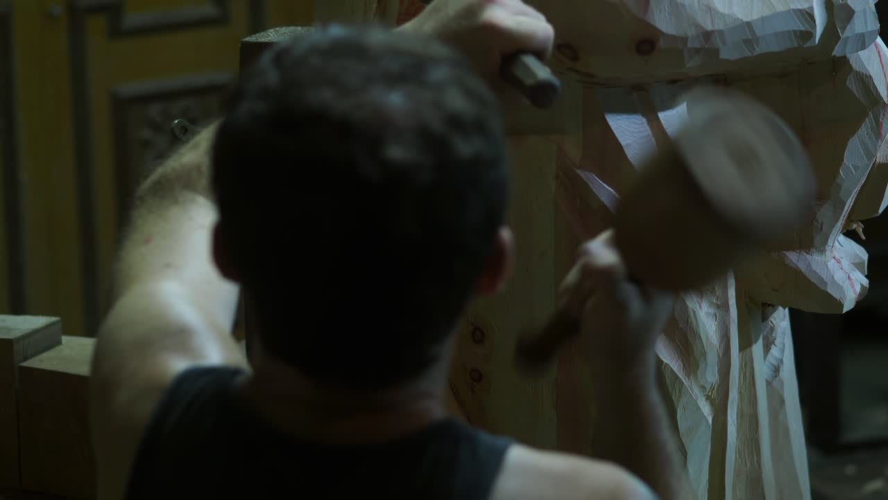 Craftsman strikes a chisel with a wooden mallet while carving details into a large wooden statue of the Virgin and Child, focusing on precision work inside a dimly lit workshop