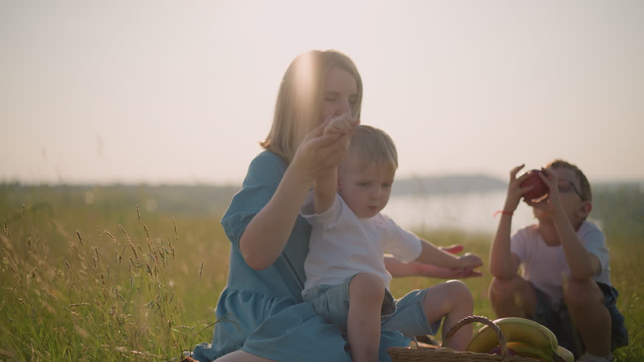 A caring mother in a blue dress with her young son on her lap, gently holding his hands as they sit in the grass. His older brother, holding an apple, in the background near a basket of fruit