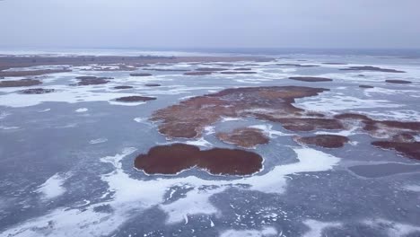 aerial view of frozen lake liepaja during the winter, blue ice with cracks, dry yellowed reed islands, overcast winter day, wide drone shot moving forward