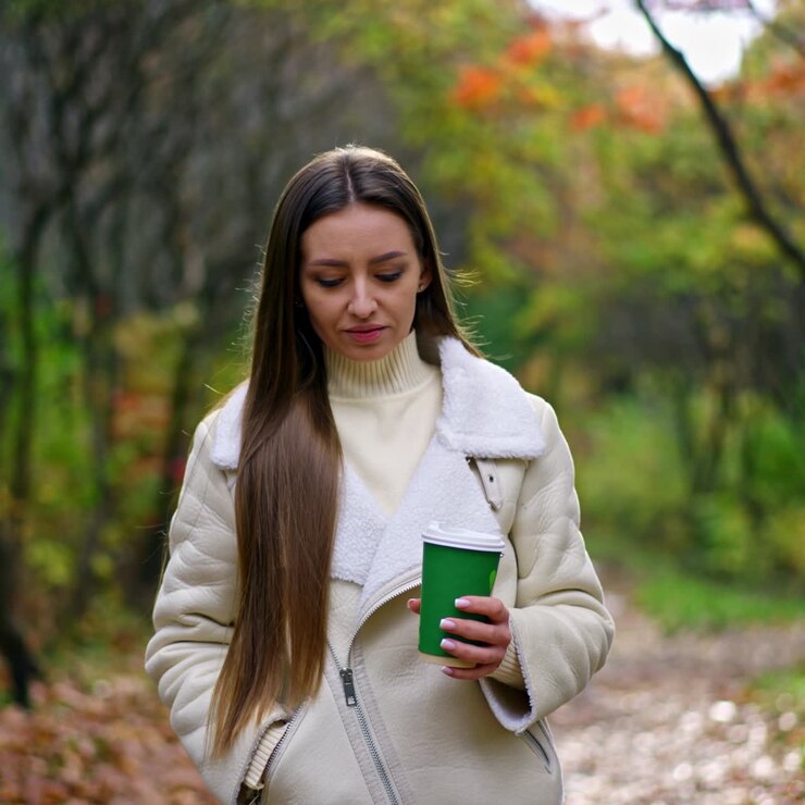 Bellissima donna dai lunghi capelli, con un bicchiere di carta in mano, passeggia nel parco. La signora si gode una calda giornata autunnale immersa nella natura. Sfondo sfocato