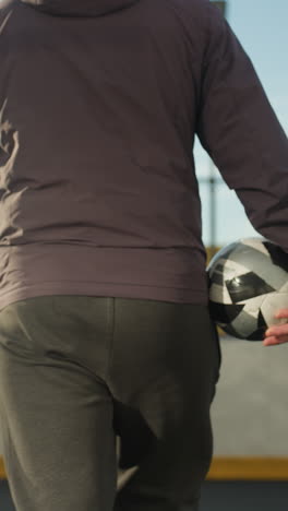 vista trasera de una persona sosteniendo una pelota de fútbol mientras entra en la arena deportiva, mostrando el entorno urbano con iluminación natural suave, el individuo está vestido con ropa deportiva casual