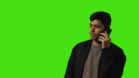 unhappy young man talking on mobile phone standing against green screen studio background