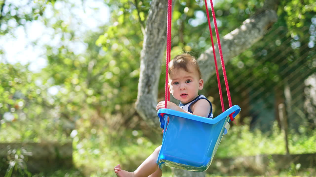 Lovely toddler swinging on a blue swing in the garden. Baby looking attentively into camera. Side view. Blurred backdrop.