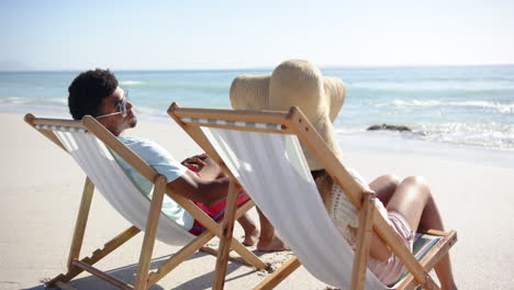 un hombre biracial y una joven mujer caucásica se relajan en sillas de playa frente al mar.