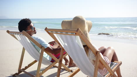 biracial man and young caucasian woman relax on beach chairs facing the sea