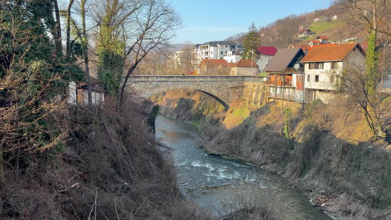 Steinbrücke über den Fluss Moravica in Ivanjica, Serbien, die ein enges Flusstal mit Häusern am Hang und fließendem Wasser überquert. Ideal für Tourismuswerbung und Videonutzung.