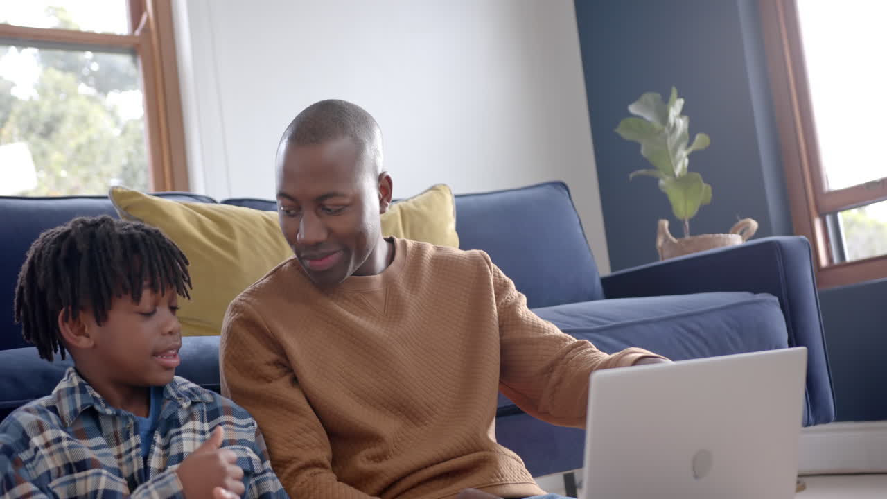 Happy african american father and son sitting on floor and using laptop at home, slow motion