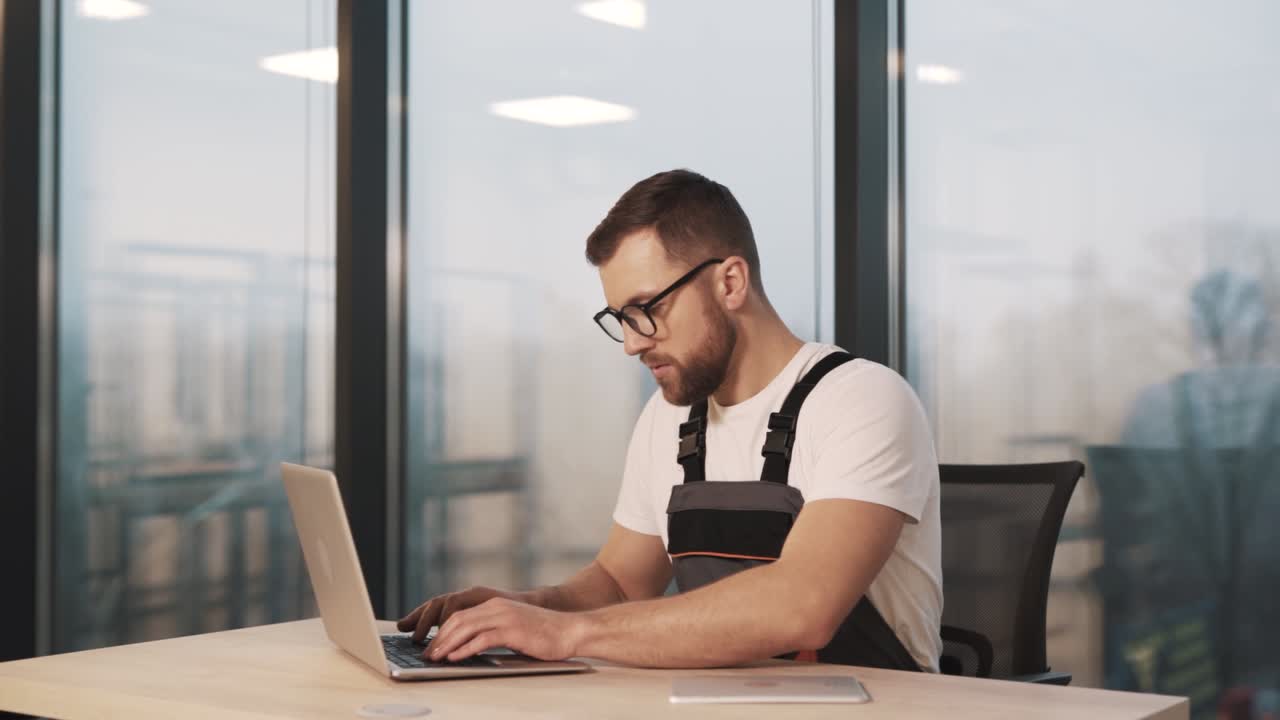 Un giovane tecnico in uniforme da lavoro e occhiali diagnostica un laptop in un ufficio moderno con un grande.