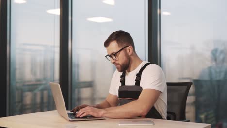 A-young-technician-in-a-work-uniform-and-glasses-diagnoses-a-laptop-in-a-modern-office-with-a-large