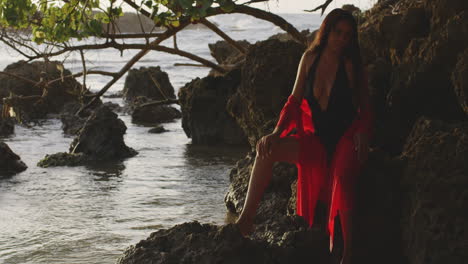 Modelo-De-Chica-Latina-En-Traje-De-Baño-Negro-Y-Rojo-Posa-Sentada-En-Las-Rocas-De-La-Playa-Tropical-Al-Amanecer,-Tiro-Panorámico