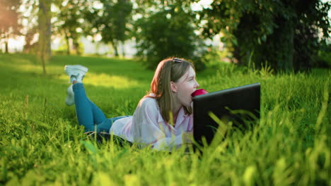 young woman lying on grass using laptop outdoors while enjoying an apple, surrounded by vibrant greenery and trees, creating a serene and natural work environment in bright daylight