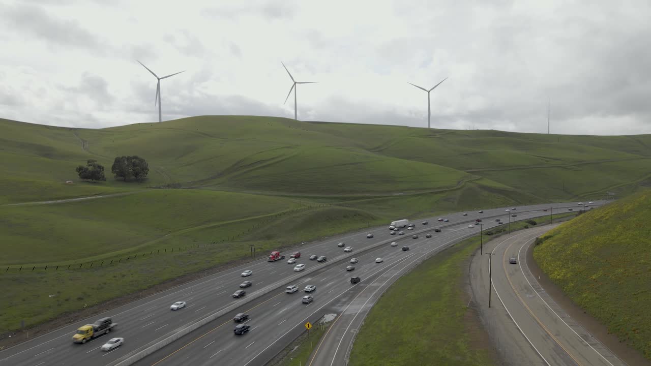 Under a canopy of clouds, the drone glides towards the green undulating terrain of the Altamont Pass, capturing Highway 580's winding journey amidst the windmills and the constant vehicular presence