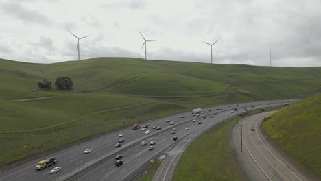 Unter-Einem-Wolkendach-Gleitet-Die-Drohne-Auf-Das-Grüne,-Hügelige-Gelände-Des-Altamont-Passes-Zu-Und-Fängt-Die-Kurvenreiche-Fahrt-Des-Highway-580-Inmitten-Der-Windmühlen-Und-Der-Ständigen-Fahrzeugpräsenz-Ein