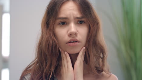 upset woman feeling sore throat and touching skin neck front bathroom mirror