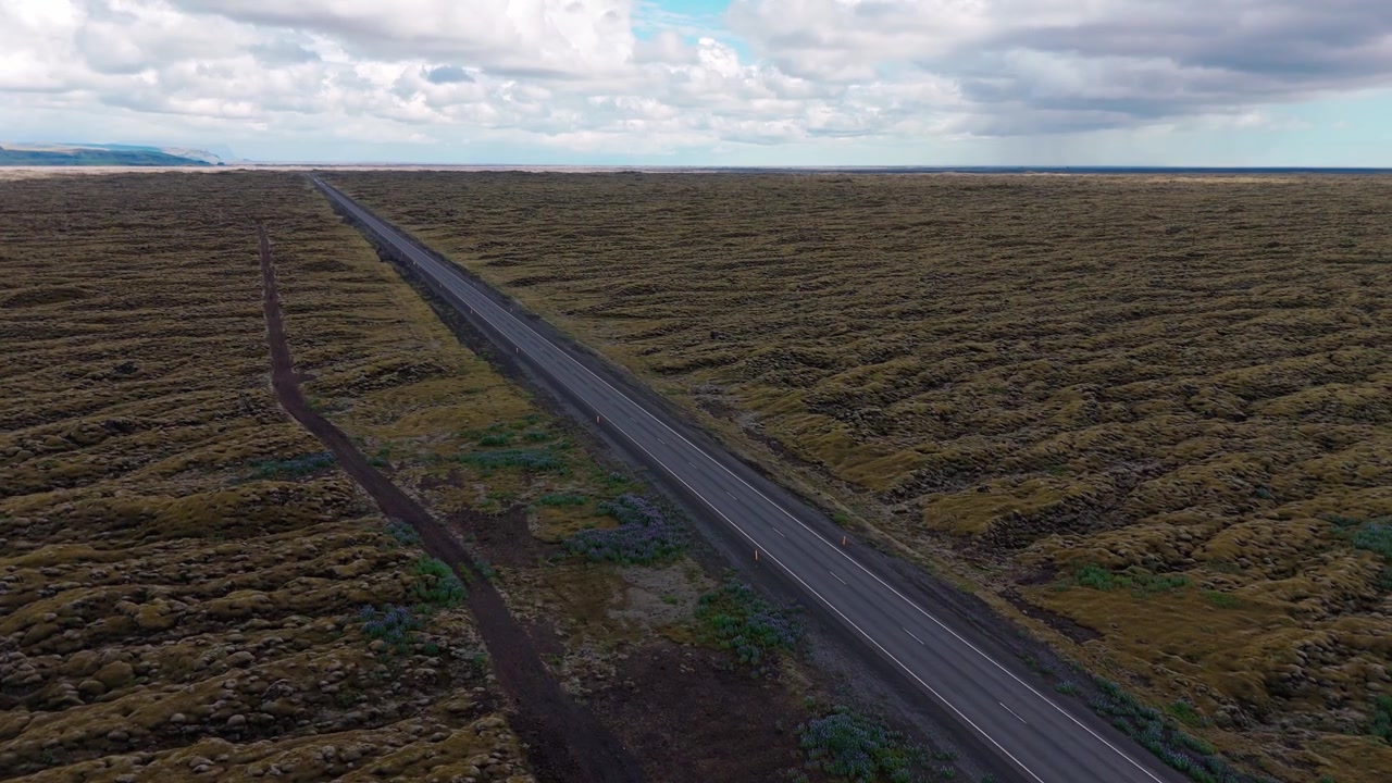 Un'ampia ripresa aerea di una strada asfaltata dritta che attraversa vasti campi di lava verdi e muschiosi sotto un cielo nuvoloso. Adatto per documentari di viaggio, sostenibilità e natura