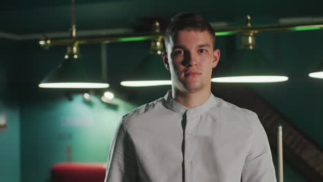 gamer in white shirt sits in focused position, holding cue stick before standing up with serious face in dimly lit pool hall. overhead lamps cast