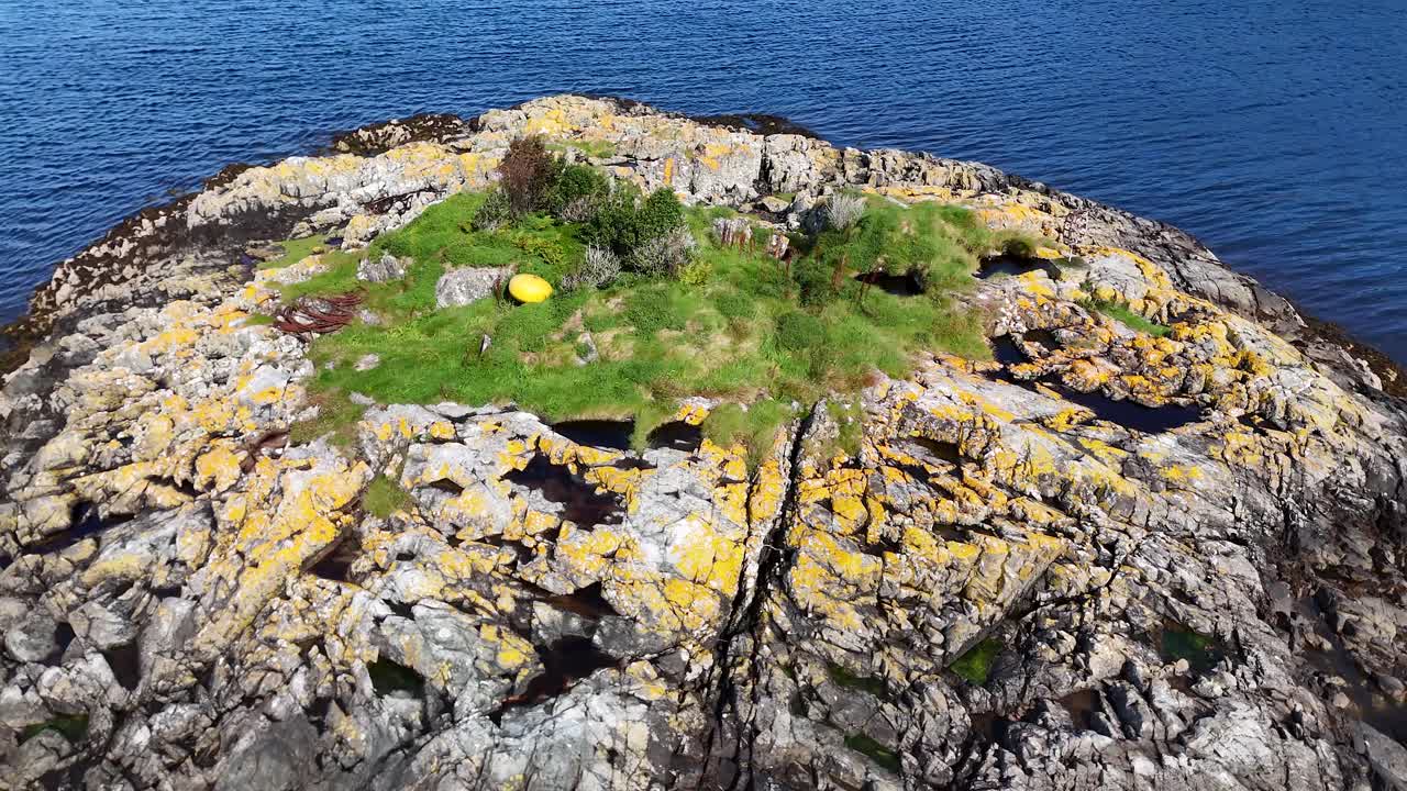 Aerial drone rises above rugged grassy coastal rocks, revealing vibrant blue water and summer textures