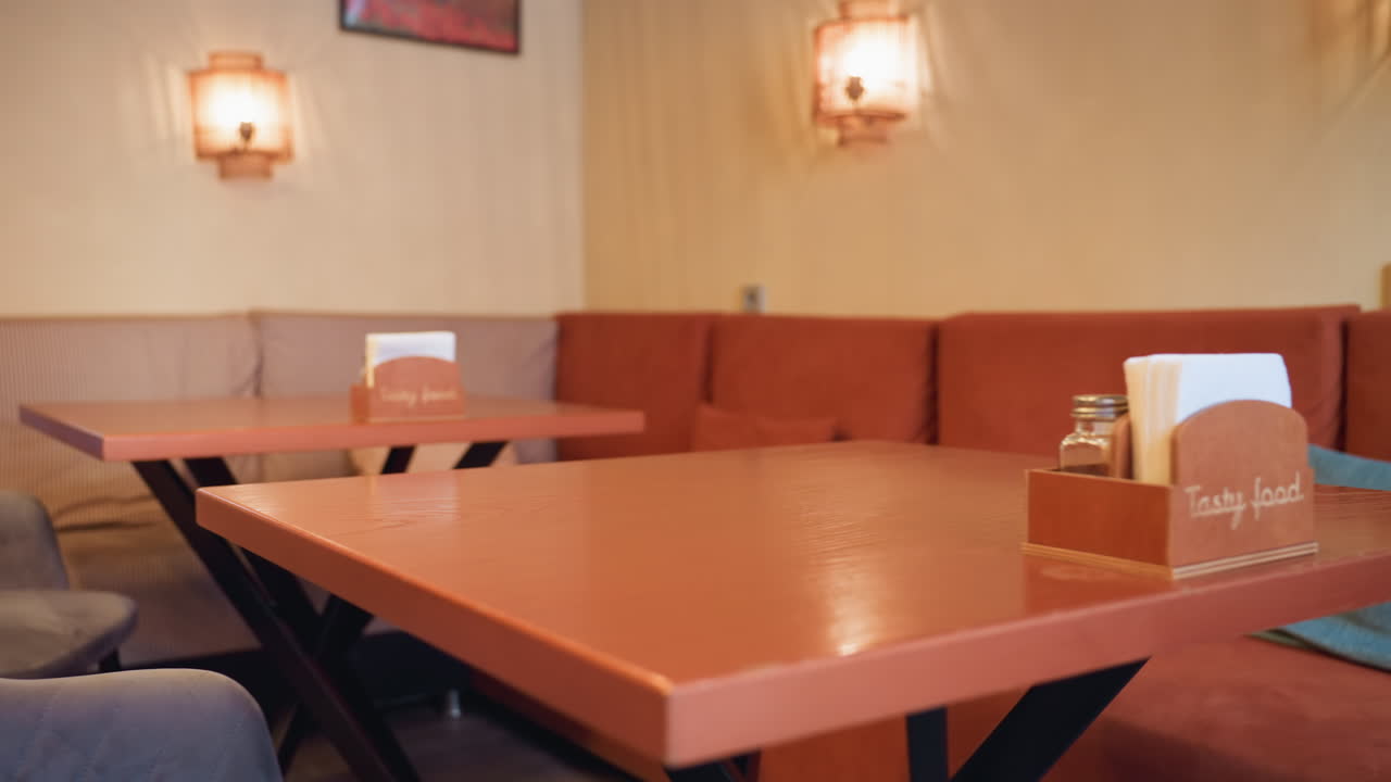 Interior display of modern restaurant showing cushioned chairs with decorative pillows, wooden tables, and warm wall lighting, creating cozy inviting atmosphere in soft toned stylish dining space