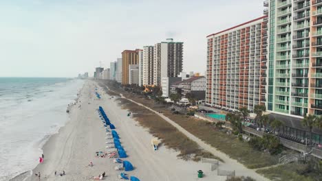 playa de arena bordeada de sombrillas azules que se extienden hacia el horizonte con gente jugando en las olas y varios edificios de gran altura en el fondo