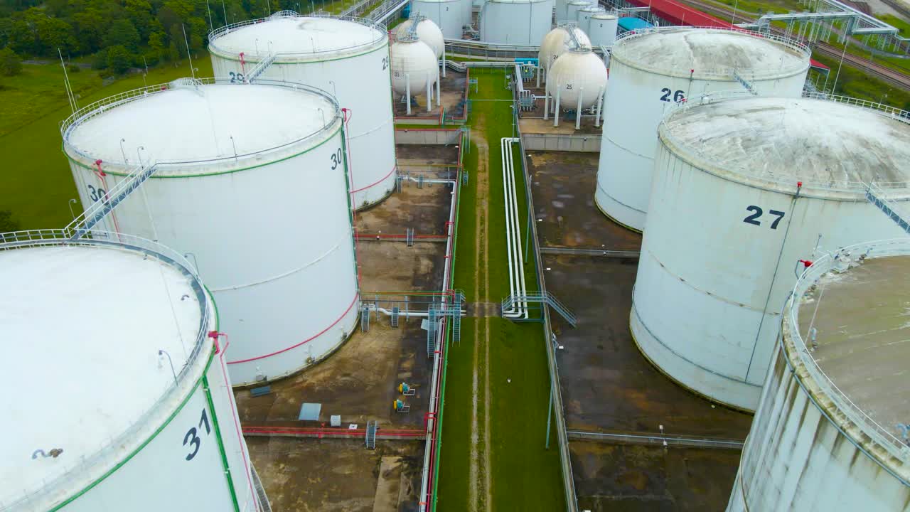 Aerial drone revealing shot footage flying over backwards above large white storage tanks in a seaside industrial harbor or port area that contain oil or gas. Small and large containers visible