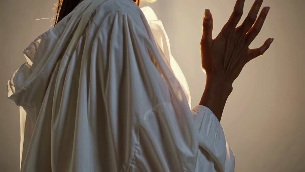 A dramatic, low-angle video shot captures a person in a flowing white garment, hands raised, bathed