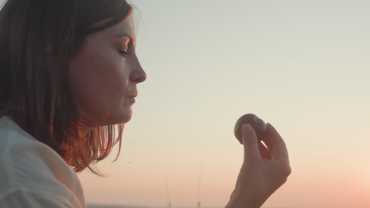 Side view of elegant woman holding ripe plum close to mouth, admiring glossy fruit against warm sunset sky, soft sunlight illuminating profile and blurred grassy field background