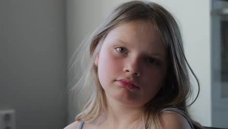 young girl with a neutral expression looks toward the camera in soft lighting indoors