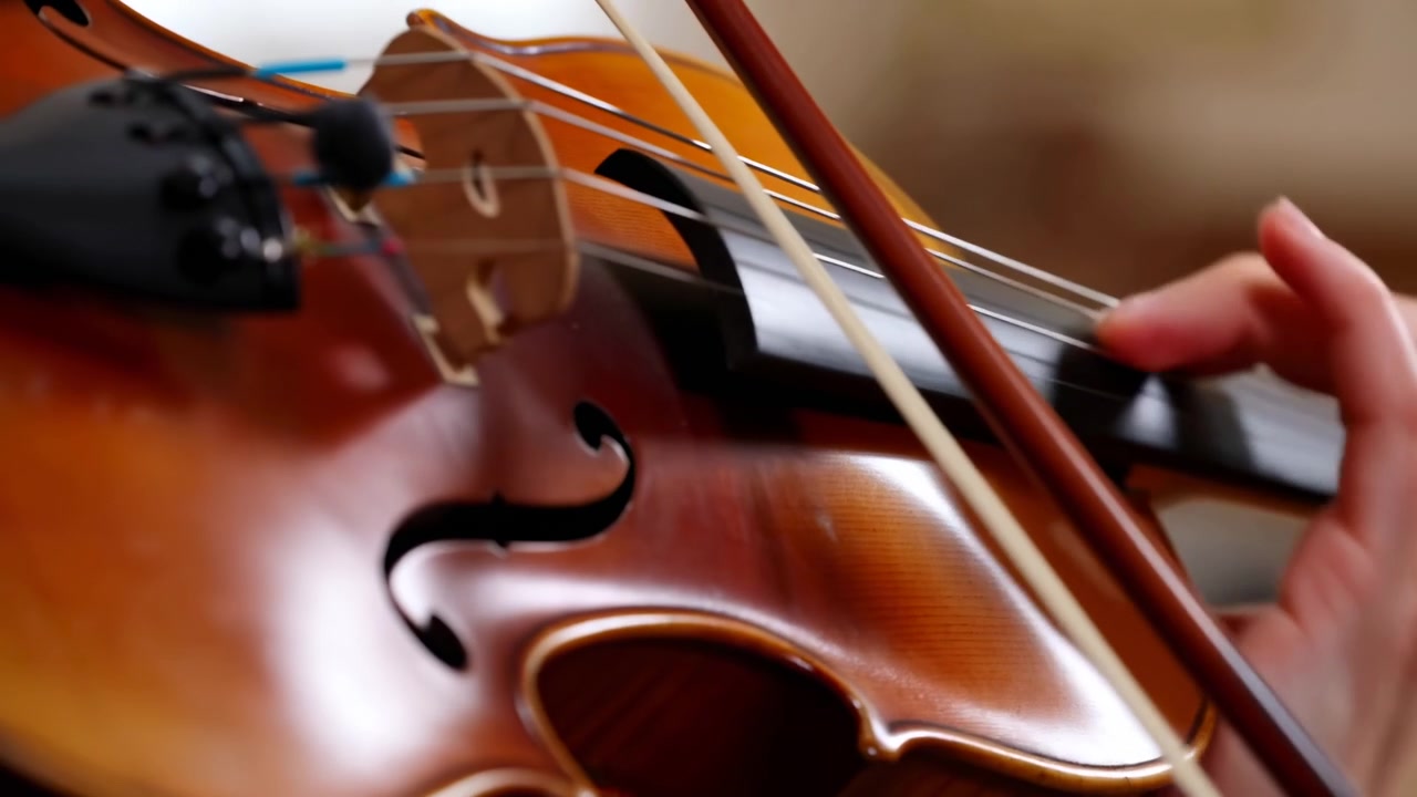 Bow glides diagonally over violin strings while one hand changes finger positions along dark fingerboard, highlighting glossy wood grain, bridge details, and warm shallow-focus setting.