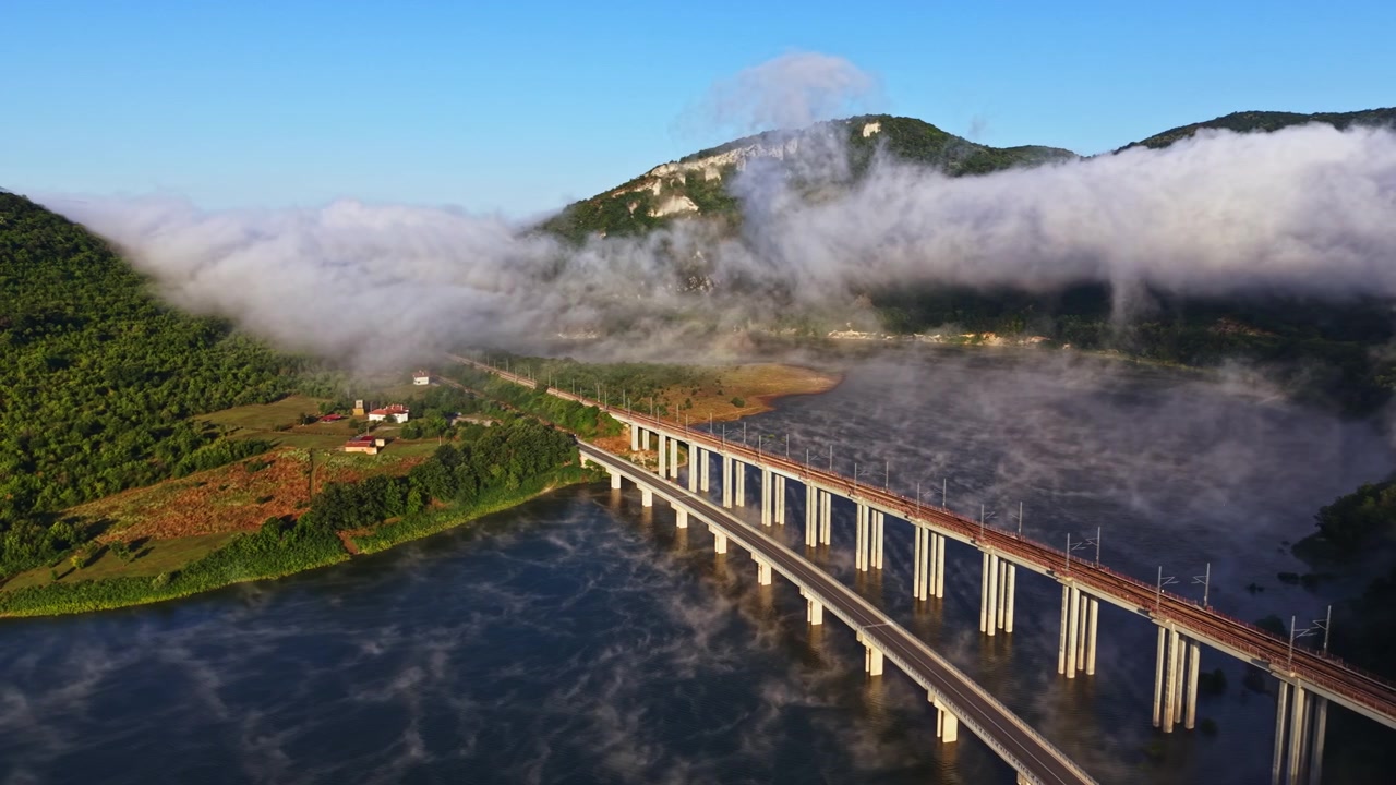 Pemandangan indah jembatan kereta api melintasi sungai dengan kabut menyelimuti pemandangan dekat gunung di pagi hari.