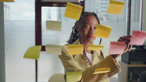 african american business woman write tasks creative ideas on sticky notes on glass board female corporate leader planning project on post it sticky notes organize work on stickers