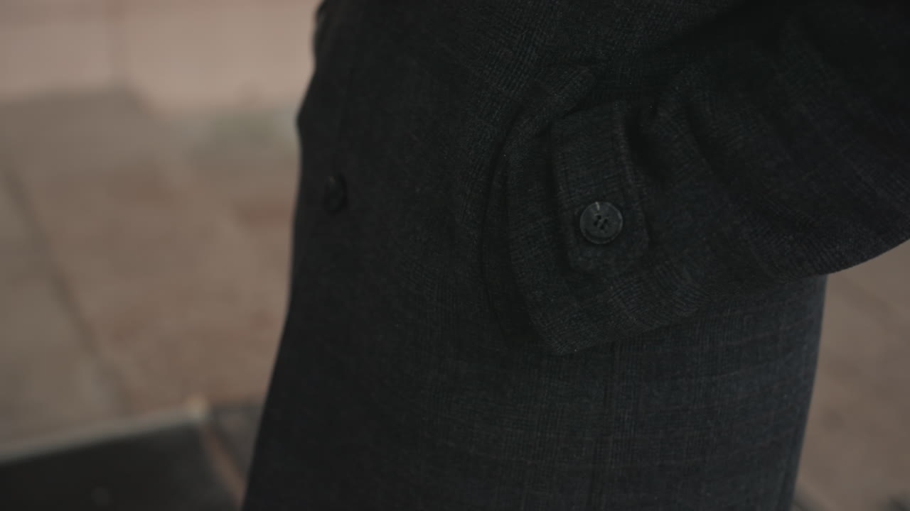 Closeup black coat pocket showing button and textured wool fabric, commuter adjusts sleeve on urban sidewalk muted tones, cinematic lighting emphasize tailoring, cuff and subtle gesture
