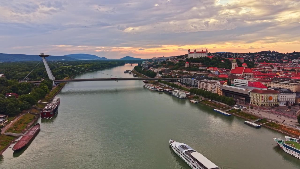 Udsigt over Donau-floden og Bratislavas bybillede under solnedgang med både og vartegn i baggrunden.