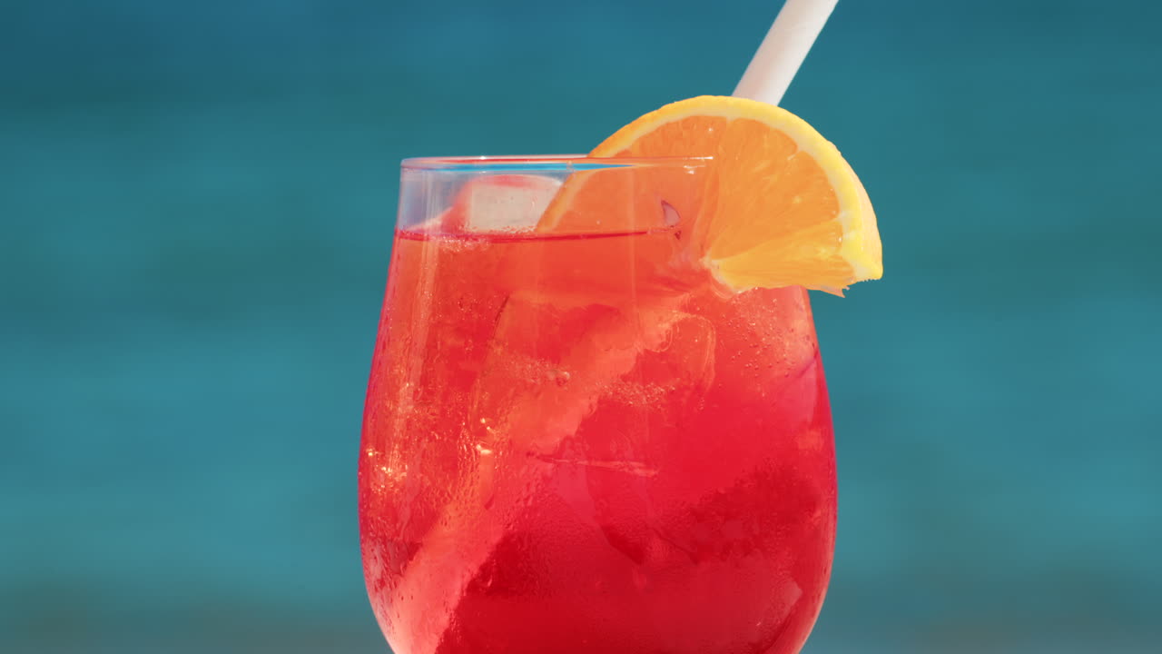 Refreshing red orange cocktail with ice and orange slice against the deep blue Mediterranean Sea
