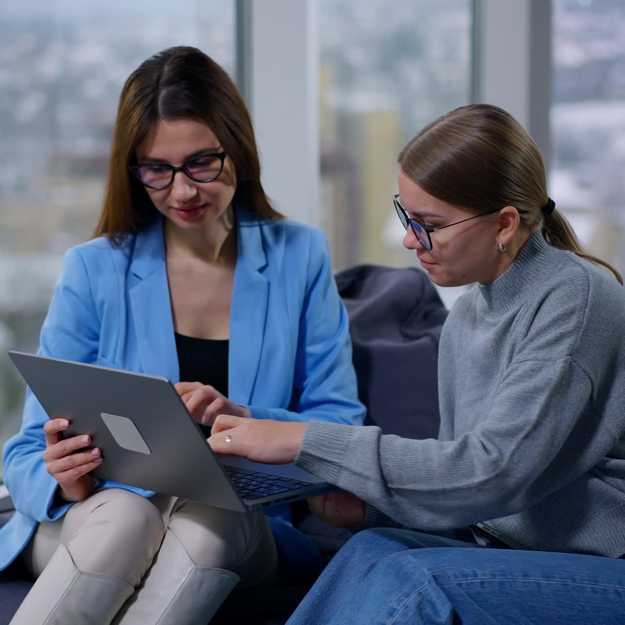 Two female employees working on some issue looking attentively on the laptop screen. Ladies work on computer communicating, sitting in soft chairs