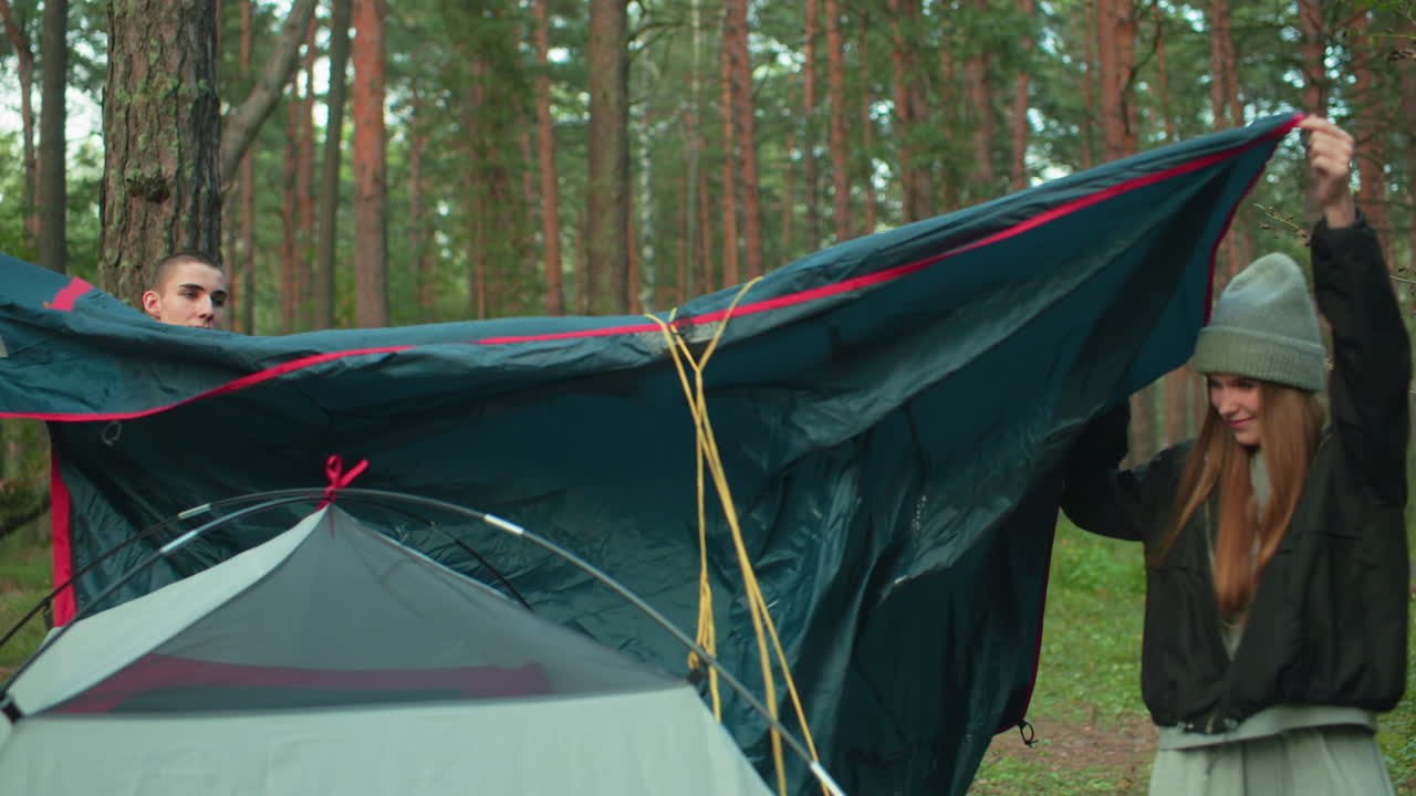Brother and sister work together to place tent cover over pitched tent in forest, sharing task with focus and care amid tall trees and lush greenery, preparing cozy shelter