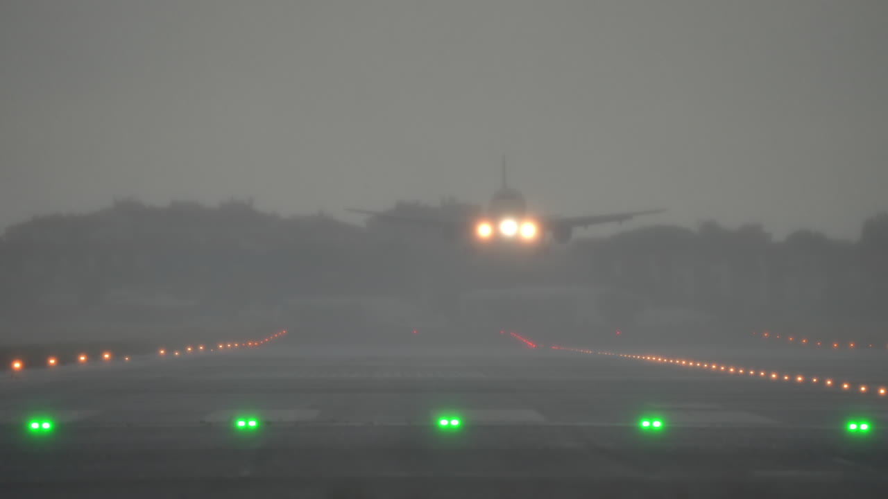 Airplane approaches runway with green and red lights visible, surrounded by fog, during heavy rain