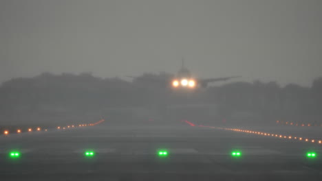 airplane approaches runway with green and red lights visible, surrounded by fog, during heavy rain