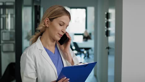 Smiling-young-female-doctor-talking-by-mobile-phone-at-the-corridor.