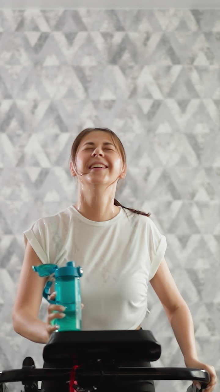 mujer riendo nerviosamente con botella en la cinta de correr en casa. mujer atlética alegre corre con el cabello mojado en el entrenador en el interior. tiempo de entrenamiento inspirador en la casa