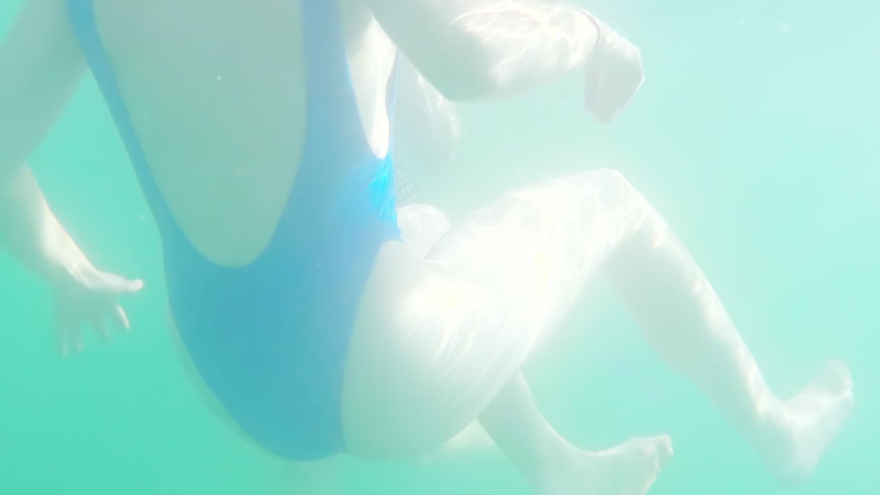 In a blue swimsuit, a woman swims in the summer water sun. Rays of light shine through the surface of the water onto her body.