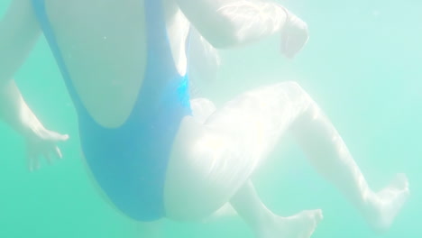 in a blue swimsuit, a woman swims in the summer water sun. rays of light shine through the surface of the water onto her body.