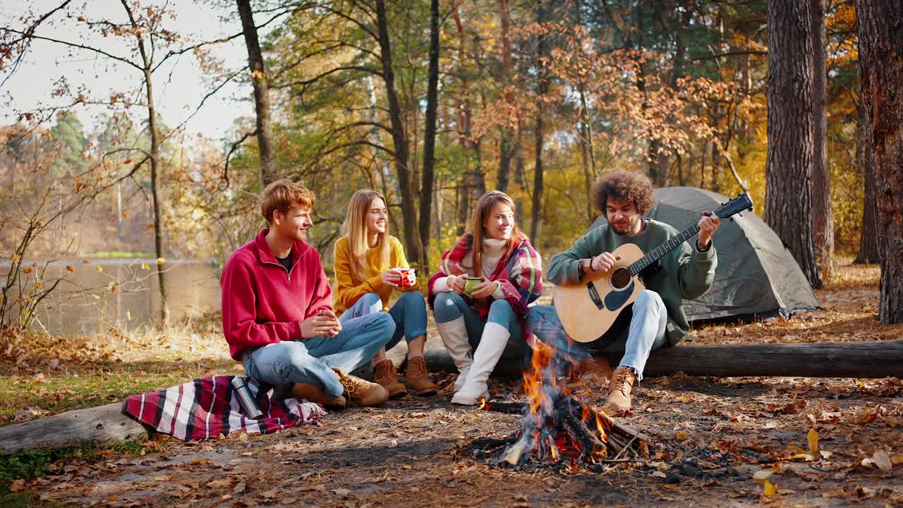 amigos sonriendo, tocando la guitarra y escuchando la música mientras están sentados en el tronco en la madera de otoño cerca del río. fuego de campamento ardiente. cámara lenta